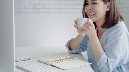 Beautiful young smiling woman working on laptop while enjoying drinking warm coffee sitting in a living room at home. Enjoying time at home. Asian business woman working in her home office.の写真素材