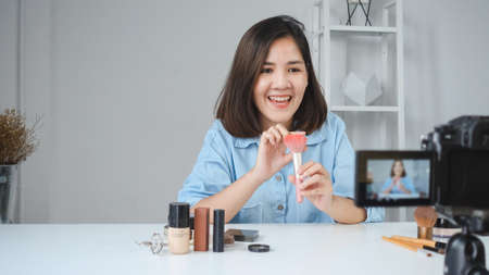 Happy smiling asian woman or beauty blogger with brush and camera recording video and waving hand at home. Beauty videoblog blogging people concept.の写真素材