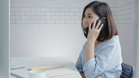 Beautiful young smiling asian woman working on laptop while sitting in a living room at home. Asian business woman using phone for work in her home office. Enjoying time at home.の写真素材