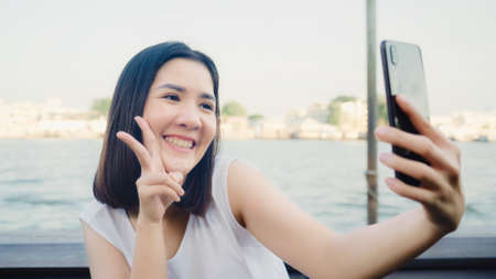 Traveler Asian blogger woman clicking selfie using mobile at cafe, beautiful female using smart phone make vlog and live in social media while enjoying the journey holiday travel near river in city.の写真素材