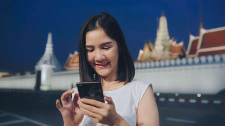 Backpacker Asian woman travel in Bangkok, Thailand. Young Asian tourist girl using mobile phone checking social media at night while walk along the street enjoying the journey holiday travel concept.の写真素材