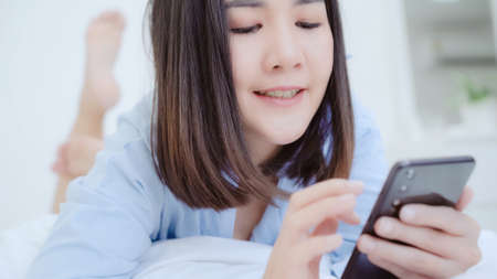 Young Asian woman using smartphone while lying on bed after wake up in the morning, Beautiful attractive Japanese girl smiling relax in bedroom at home. Enjoying time lifestyle women at home concept.の写真素材