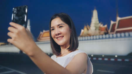 Happy young Asian girl tourist with casual style hold mobile phone take selfie photo peaceful picture of background temple in bangkok city at night, Lifestyle tourist travel holiday concept.の写真素材