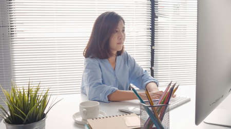 Beautiful young smiling Asian woman working on laptop while at home in office work space. Businesswoman working from home via portable computer writing on keyboard. Enjoying time at home.の写真素材