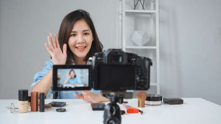 Happy smiling asian woman or beauty blogger with brush and camera recording video and waving hand at home. Beauty videoblog blogging people concept.の写真素材