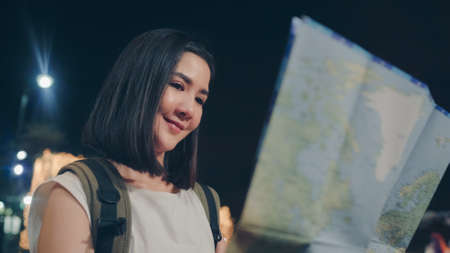 Backpacker Asian woman travel in Bangkok, Thailand. Young Asian tourist girl looking maps for direction places at night while walk along the street enjoying the journey holiday travel concept.の写真素材