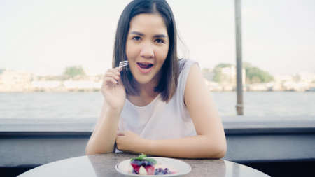 Blogger Asian friendly woman influencer eating cake at cafe. Young lady happy relax fun using technology mobile phone record vlog video upload in social media holiday travel near river in city.の写真素材