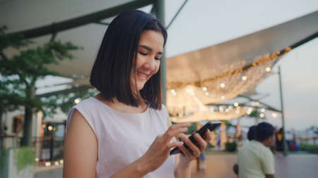 Happy young Asia woman blogger tourist with casual style hold smartphone slide screen on mobile phone with river view sunset near cafe in city town at night. Lifestyle tourist travel holiday concept.の写真素材