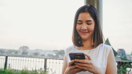 Happy young Asian woman blogger tourist with casual style hold smartphone look with smile joyful with beautiful sunset on river view near cafe in city town. Lifestyle tourist travel holiday concept.の写真素材
