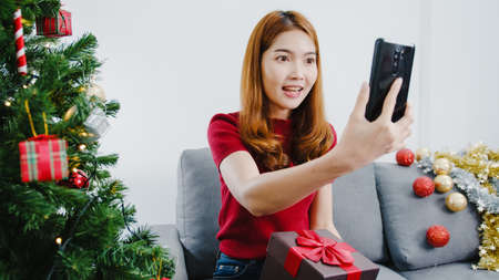 Young Asian female using smart phone video call talking with couple with X'Mas present box, Christmas tree decorated with ornament in living room at home. Christmas and New Year holiday festival.の写真素材