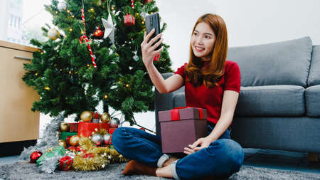 Young Asian female using smart phone video call talking with couple with X'Mas present box, Christmas tree decorated with ornament in living room at home. Christmas and New Year holiday festival.の写真素材