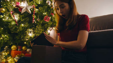 Young Asia female having fun opening X'Mas present box near Christmas tree decorated with ornaments in living room at home. Merry Christmas night and Happy New Year holiday festival.の写真素材
