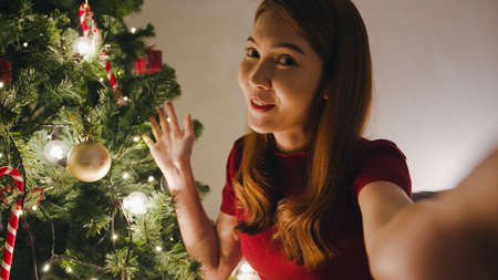Young Asia female using smart phone video call talking with couple, Christmas tree decorated with ornament in living room at home. Social distancing, Christmas night and New Year holiday festival.の写真素材