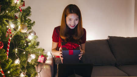 Young Asia female using tablet video call talking with couple with X'Mas present box, Christmas tree decorated with ornament in living room at home. Christmas night and New Year holiday festival.の写真素材