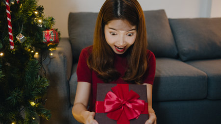 Young Asia female having fun opening X'Mas present box near Christmas tree decorated with ornaments in living room at home. Merry Christmas night and Happy New Year holiday festival.の写真素材