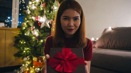 Young Asia female using mobile phone video call talking with couple with X'Mas present box, Christmas tree decorated with ornament in living room at home. Christmas night and New Year holiday festivalの写真素材