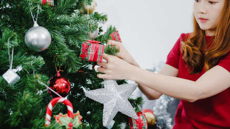 Asian female decorated with ornament on Christmas tree at Christmas and New Year festival in living room at home. Xmas celebration event preparation or winter holidays festival indoor party concept.の写真素材
