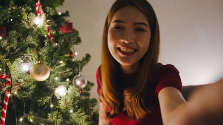 Young Asia female using smart phone video call talking with couple, Christmas tree decorated with ornament in living room at home. Social distancing, Christmas night and New Year holiday festival.の写真素材