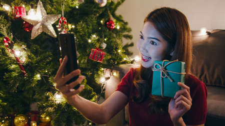 Young Asia female using smart phone video call talking with couple with X'Mas present box, Christmas tree decorated with ornament in living room at home. Christmas night and New Year holiday festival.の写真素材