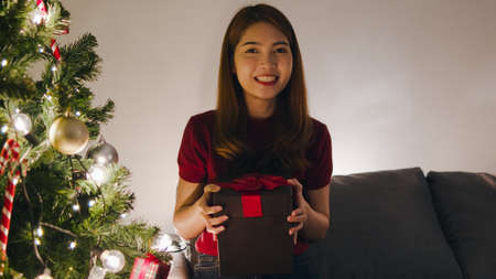 Young Asia female using mobile phone video call talking with couple with X'Mas present box, Christmas tree decorated with ornament in living room at home. Christmas night and New Year holiday festivalの写真素材