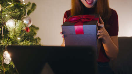 Young Asia female using tablet video call talking with couple with X'Mas present box, Christmas tree decorated with ornament in living room at home. Christmas night and New Year holiday festival.の写真素材