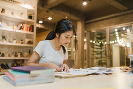 Asian student women reading books in library at university. Young undergraduate girl do homework, read textbook, study hard for knowledge on lecture desk at college campus overtime night.の写真素材