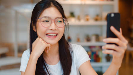 Blogger Asian friendly women influencer waving hand making video call at night cafe. Young lady happy relax fun using technology mobile phone record vlog upload in social media at college campus.の写真素材