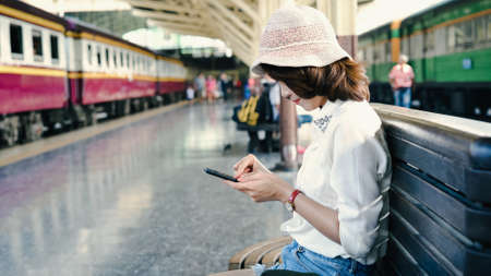 Traveler backpacker Asian woman travel in Bangkok, Thailand. Happy young female sitting on bench using smartphone for talking, reading and texting at train station.の写真素材