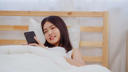Young Asian woman using smartphone checking social media feeling happy smiling while lying on bed after wake up in the morning, Beautiful attractive Japanese girl smiling relax in bedroom at home.の写真素材