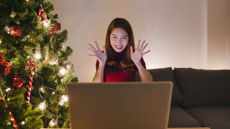 Young Asia female using laptop video call talking with couple with X'Mas present box, Christmas tree decorated with ornament in living room at home. Christmas night and New Year holiday festival.の写真素材