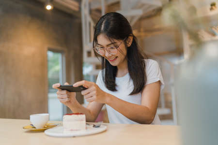 Blogger Asian friendly women influencer eat cake at night cafe. Beautiful young lady happy relax fun using technology mobile phone taking a photo of her food upload in social media at college campus.の写真素材