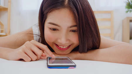 Young Asian woman using smartphone checking social media feeling happy smiling while lying on bed after wake up in the morning, Beautiful attractive Japanese girl smiling relax in bedroom at home.の写真素材