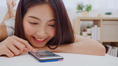Young Asian woman using smartphone checking social media feeling happy smiling while lying on bed after wake up in the morning, Beautiful attractive Japanese girl smiling relax in bedroom at home.の写真素材