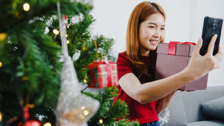 Young Asian female using smart phone video call talking with couple with X'Mas present box, Christmas tree decorated with ornament in living room at home. Christmas and New Year holiday festival.の写真素材