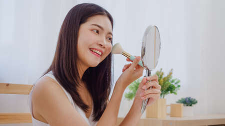 Asian woman using powder by brush make up in front mirror, Happy female using beauty cosmetics to improve herself ready to working in bedroom at home. Lifestyle women at home concept.の写真素材