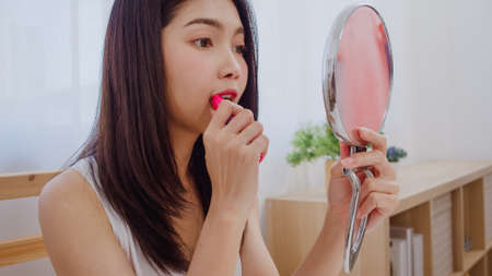 Young Asian woman using lipstick make up in front mirror, Happy female using beauty cosmetics to improve herself ready to working in bedroom at home. Lifestyle women at home concept.の写真素材