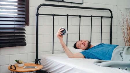 Young Asian man wake up in the morning, Asia male turn off alarm clock after awake on bed in bedroom at home. Handsome men nap, sleepy relax in modern house concept.の写真素材
