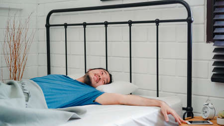 Young Asian man wake up in the morning, Asia male turn off alarm clock in mobile phone after awake on bed in bedroom at home. Handsome men nap, sleepy relax in modern house concept.の写真素材
