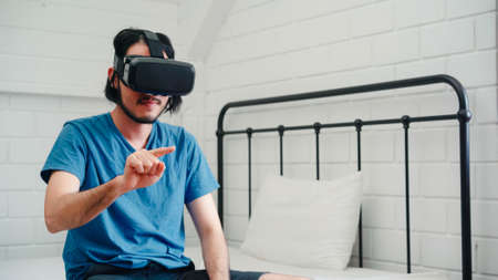 Young Asian man using glasses virtual reality simulator playing video game in bedroom, Japanese male feeling happy using relax time lying on bed at home. Teen play games relax at home concept.の写真素材