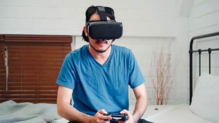 Asian man using glasses virtual reality simulator and joystick playing racing game in bedroom, Japanese male feeling happy using relax time lying on bed at home. Teen play games relax at home concept.の写真素材