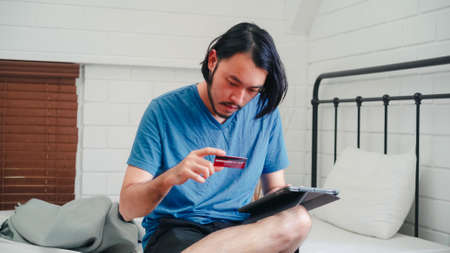 Young Asian man using tablet and credit card shopping ecommerce, male relax feeling happy online shopping sitting on bed in bedroom at home. Teen men shop in internet, online communication concept.の写真素材