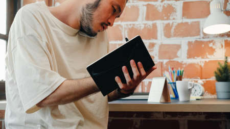 Freelance Asia guy working at his desk in office taking a call on his mobile phone while writing notes on a notepad. Working from home, remotely work, social distancing, quarantineの写真素材