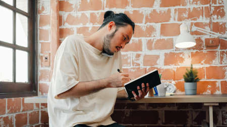 Freelance Asia guy working at his desk in office taking a call on his mobile phone while writing notes on a notepad. Working from home, remotely work, social distancing, quarantineの写真素材