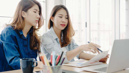 Attractive smart Asian business woman in smart casual wear working on laptop while sitting on desk while her angry manager standing angry throwing papers on office desk. Women work at office concept.の写真素材