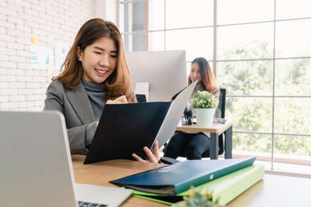 Attractive smart Asian business woman in smart casual wear working on laptop while sitting on desk while her angry manager standing angry throwing papers on office desk. Women work at office concept.の写真素材