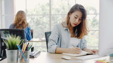 Attractive smart Asian business woman in smart casual wear working on laptop while sitting on desk while her angry manager standing angry throwing papers on office desk. Women work at office concept.の写真素材