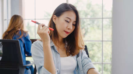 Attractive smart Asian business woman in smart casual wear working on laptop while sitting on desk while her angry manager standing angry throwing papers on office desk. Women work at office concept.の写真素材
