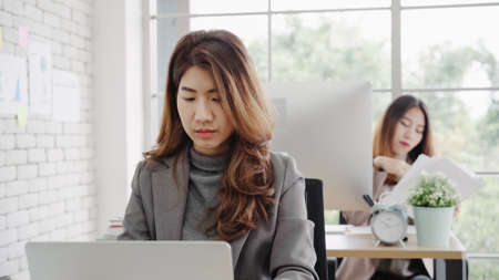 Attractive smart Asian business woman in smart casual wear working on laptop while sitting on desk while her angry manager standing angry throwing papers on office desk. Women work at office concept.の写真素材
