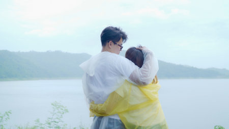 Young Asian sweet couple feeling happy using romantic time playing rain while wearing raincoat standing near lake. Lifestyle couple enjoy and relax in rainy day.の写真素材