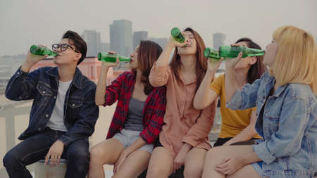 Group of young asian women and man people dancing and raising their arms up in air to the music played by dj at sunset urban party on rooftop. Young asian girls and boy friends hanging out with drinksの写真素材
